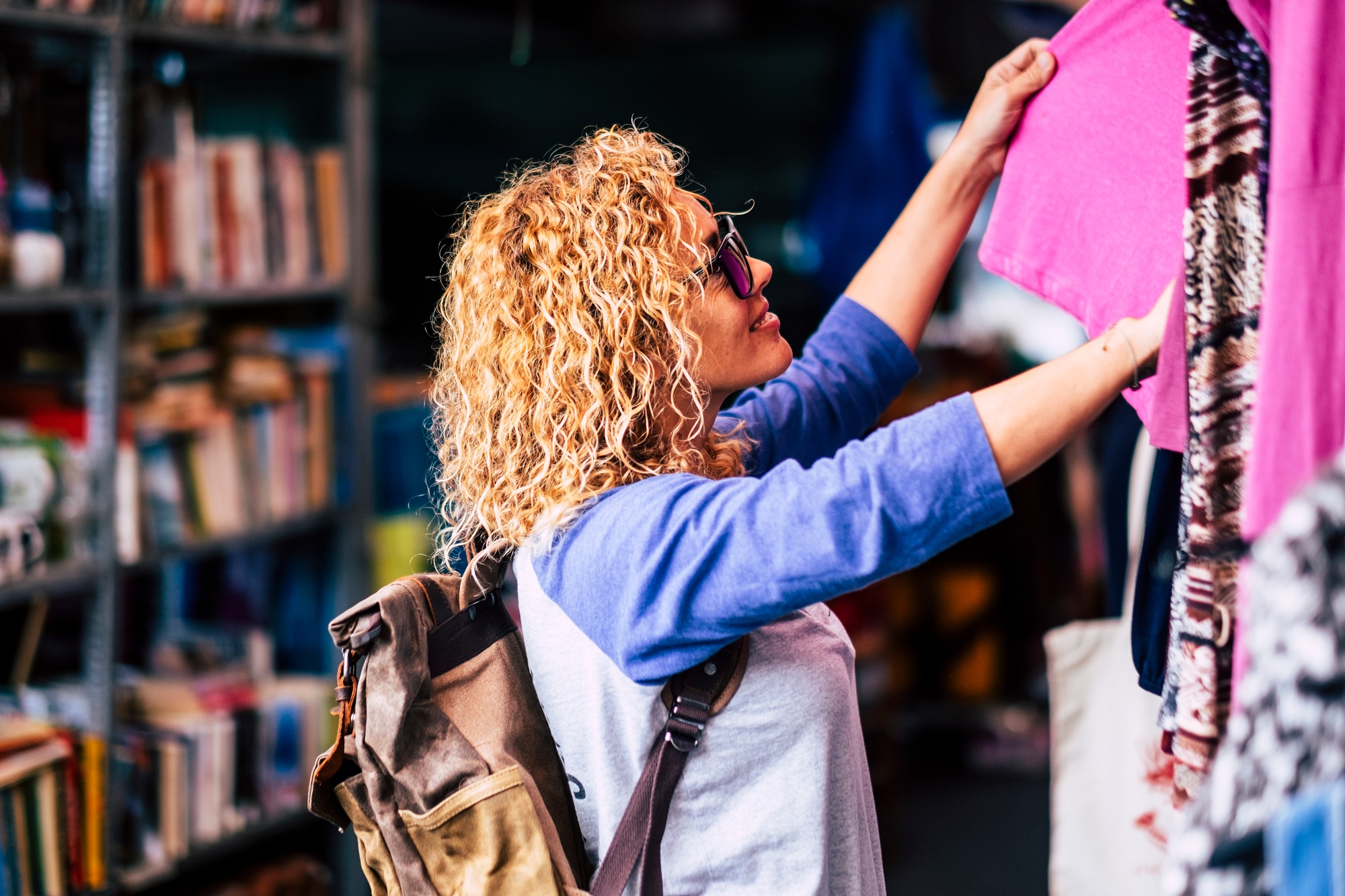 Cheerful traveler young blonde curly woman looking and choosing clothes at the used flea market